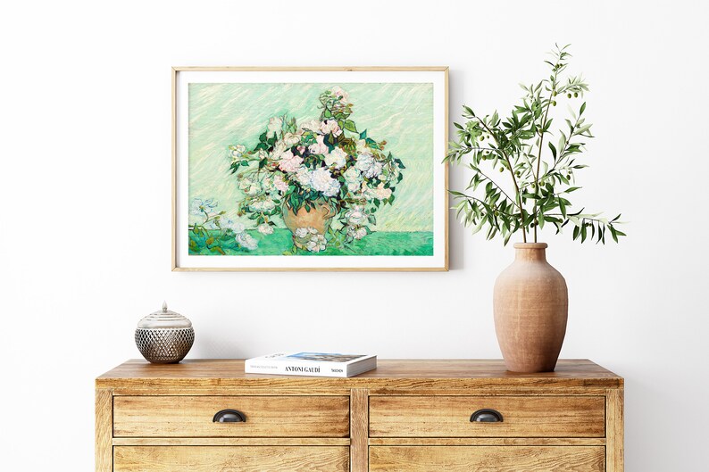 K&ouml;nnte beinhalten: Ein gerahmtes Gem&auml;lde mit einer Vase voller wei&szlig;er Rosen. Das Gem&auml;lde ist in einem hellen Holzrahmen gefasst und h&auml;ngt an einer wei&szlig;en Wand. Die Wand ist au&szlig;erdem mit einer Vase mit gr&uuml;nen Bl&auml;ttern und einer kleinen dekorativen Schale verziert.