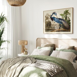 K&ouml;nnte beinhalten: Ein gr&uuml;nes und beiges Schlafzimmer mit einem Bett, das mit einer gr&uuml;nen Bettdecke und einer grauen Wolldecke ausgestattet ist. Ein gerahmtes Bild eines blauen Reihers h&auml;ngt an der Wand &uuml;ber dem Bett. Ein Kopfteil aus Weidengeflecht und eine Weidenlampe sind ebenfalls zu sehen.