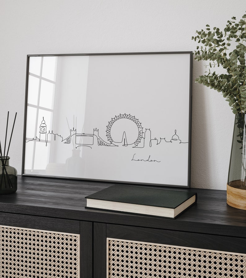May include: Black and white line art print of the London skyline, including the London Eye, Big Ben, and Tower Bridge. The print is framed in a black frame and is hanging on a wall above a black cabinet with woven cane doors.