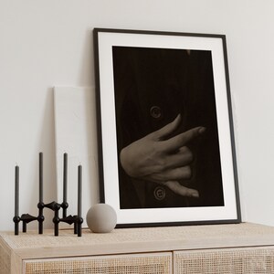 Op de afbeelding: Een zwart ingelijste foto van een hand met een donkere achtergrond. De foto staat op een licht houten kast met een geweven rotan voorzijde.