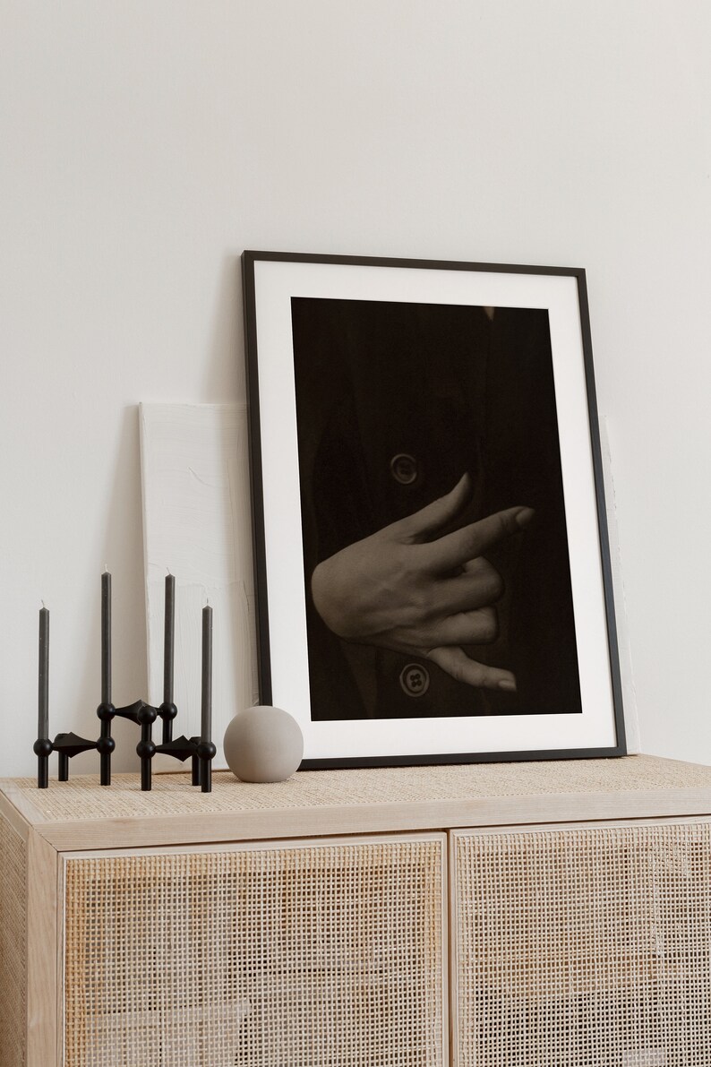 Op de afbeelding: Een zwart ingelijste foto van een hand met een donkere achtergrond. De foto staat op een licht houten kast met een geweven rotan voorzijde.