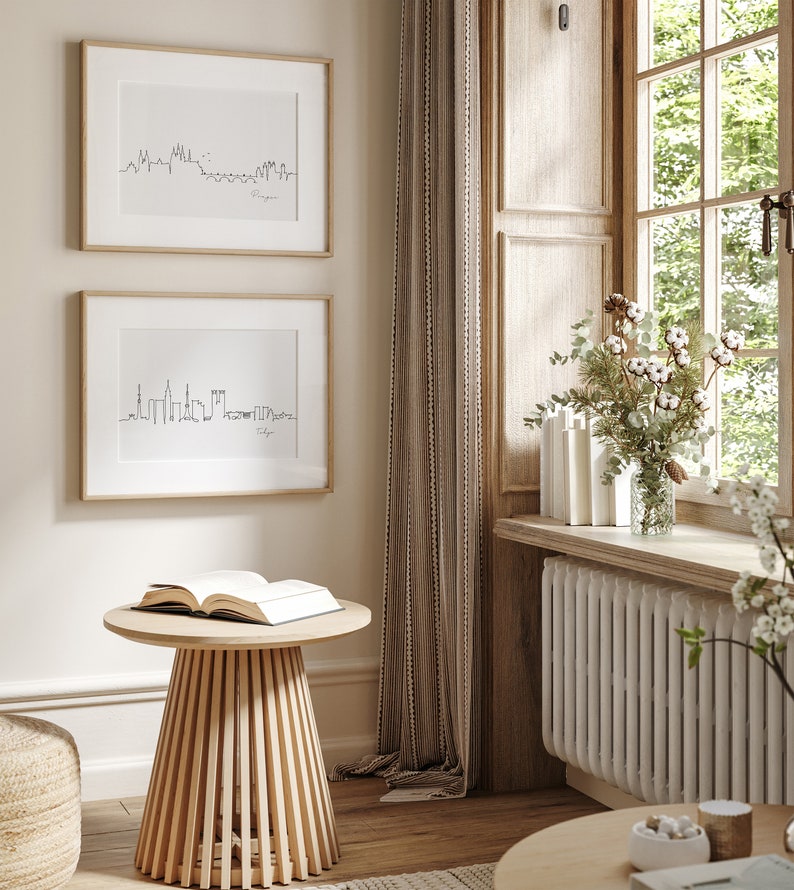 May include: Two framed black and white skyline prints, labelled "Prague" and "Tokyo", hang on a beige wall. A small, light wood side table with an open book sits below. The room has a window with a floral arrangement.