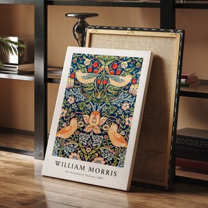 Op de afbeelding: Een canvas print van William Morris' ontwerp "The Strawberry Thieves". De print heeft een blauwe achtergrond met een herhalend patroon van vogels, bloemen en bladeren. De tekst "William Morris" en "The Strawberry Thieves | 1883" staat onderaan de print.