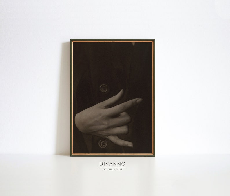Op de afbeelding: Een zwart-witfoto van een hand die een gebaar maakt. De hand is gedeeltelijk bedekt door een donker kledingstuk. De afbeelding is ingelijst in een donkergroen frame met een gouden rand. De tekst "DIVANNO ART COLLECTIVE" is onder het frame gedrukt.