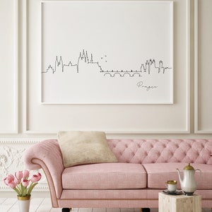 May include: A framed black and white line drawing of the Prague skyline, with the city's name in cursive. The artwork hangs above a pink tufted sofa with a beige cushion. A small wooden table holds a teapot and cup.