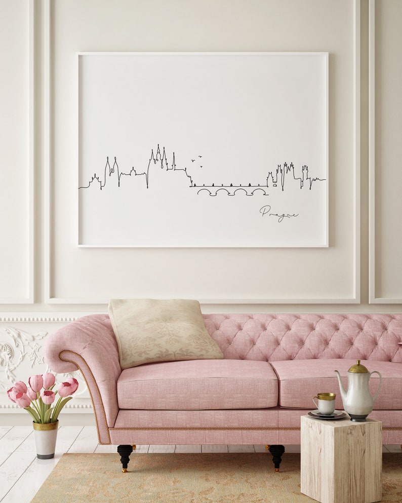 May include: A framed black and white line drawing of the Prague skyline, with the city's name in cursive. The artwork hangs above a pink tufted sofa with a beige cushion. A small wooden table holds a teapot and cup.