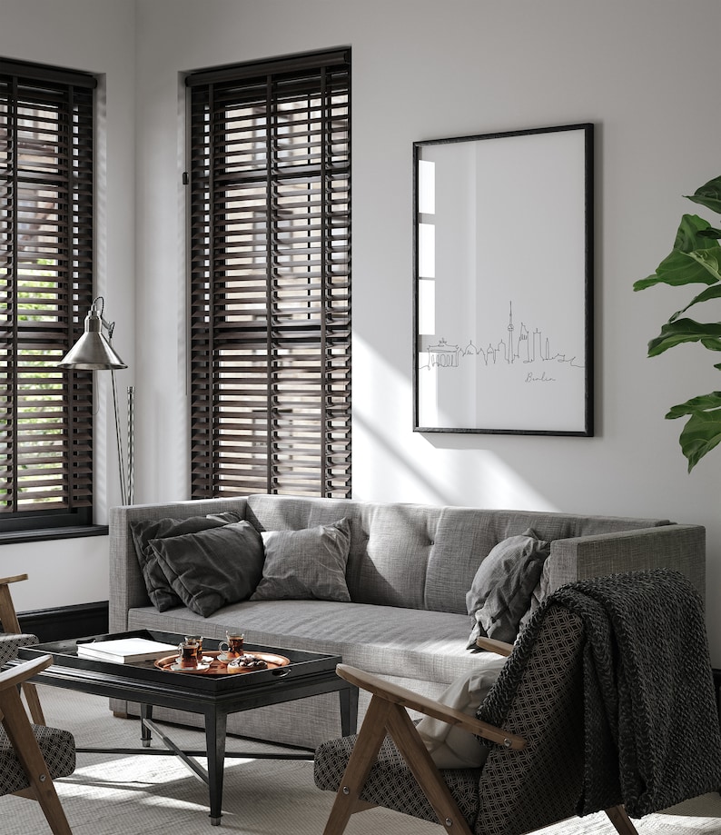 Puede incluir: Un sof&aacute; gris con almohadas negras se encuentra en una sala de estar con persianas de madera, una mesa de caf&eacute; negra y una impresi&oacute;n enmarcada del horizonte de Berl&iacute;n.