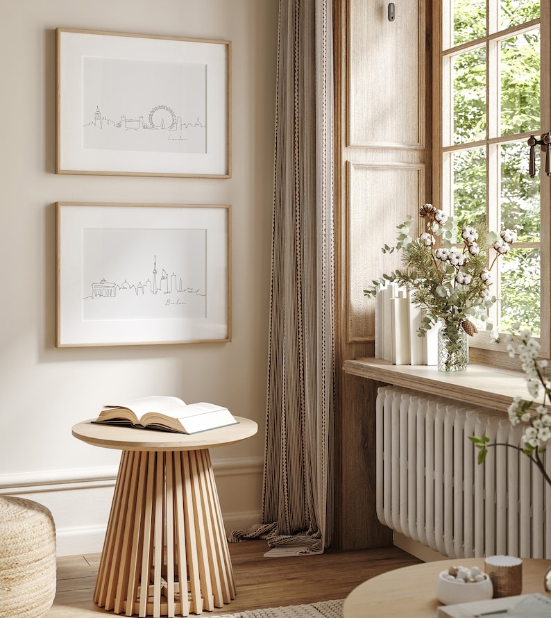 May include: Two framed black and white line drawings of city skylines. The top drawing is of London and the bottom drawing is of Berlin. The drawings are hanging on a beige wall next to a window with white wooden blinds. A wooden side table with a book on top sits in front of the drawings.