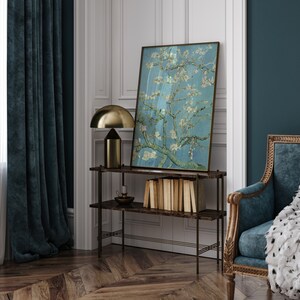Puede incluir: Una cortina de terciopelo azul oscuro cuelga frente a una ventana con cortinas blancas transparentes. Una l&aacute;mpara de mesa dorada con una pantalla en forma de hongo se encuentra sobre un estante de madera oscura con una pintura de un almendro en flor en un esquema de color azul claro y blanco. El estante forma parte de una unidad de estanter&iacute;a de marco de metal negro. Un sill&oacute;n de terciopelo azul oscuro con una manta de piel sint&eacute;tica blanca se encuentra en la esquina de la habitaci&oacute;n.