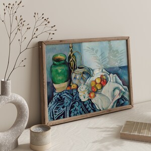 May include: A still life painting featuring a green ceramic vase, a blue and white ceramic pitcher, and a bowl of red apples. The objects are arranged on a patterned tablecloth with a white cloth draped over the side. The painting is framed in a wooden frame.