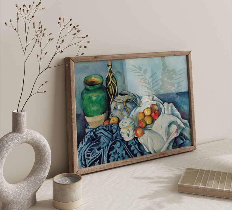 May include: A still life painting featuring a green ceramic vase, a blue and white ceramic pitcher, and a bowl of red apples. The objects are arranged on a patterned tablecloth with a white cloth draped over the side. The painting is framed in a wooden frame.