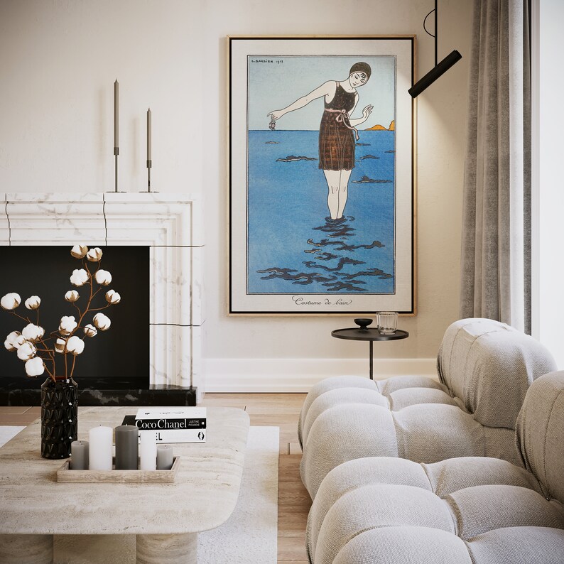 May include: A white living room with a fireplace, a white sofa, a coffee table with candles and a book, and a framed vintage poster of a woman in a swimsuit standing in the ocean. The poster reads "Costume de Bain".