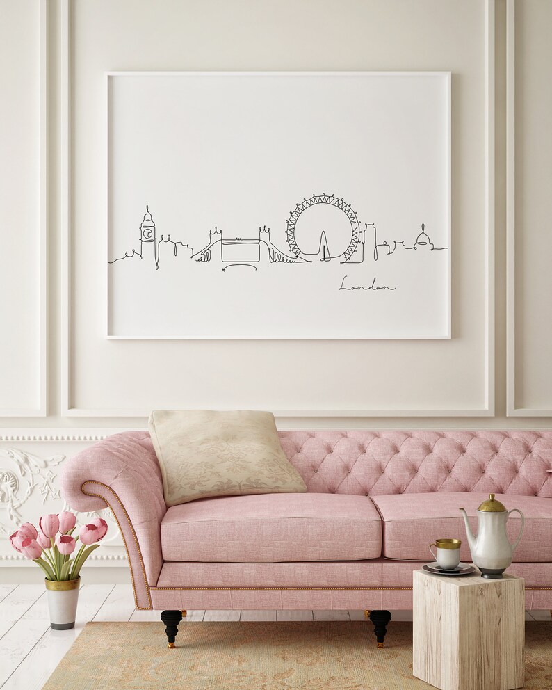 May include: A pink tufted sofa with a beige pillow in front of a white wall with a framed black and white line drawing of the London skyline, including the London Eye, Big Ben, and Tower Bridge. The text "London" is written below the skyline.