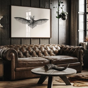 May include: A dark brown leather sofa with tufted upholstery sits in a dimly lit room. A black and white print of a bat hangs on the wall above the sofa. A decorative horn sits on a round wooden coffee table in front of the sofa.