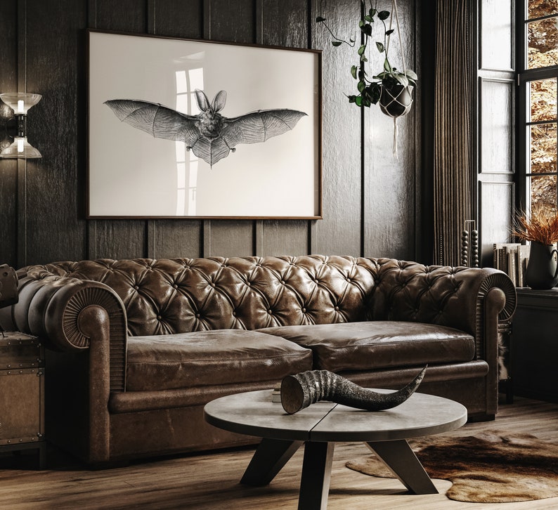 May include: A dark brown leather sofa with tufted upholstery sits in a dimly lit room. A black and white print of a bat hangs on the wall above the sofa. A decorative horn sits on a round wooden coffee table in front of the sofa.