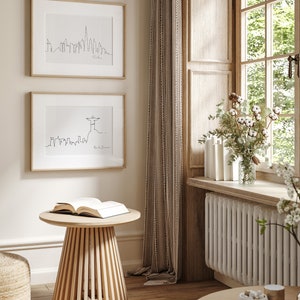 May include: Two framed black and white line drawings of city skylines, one of Dubai and one of Rio de Janeiro, hang on a beige wall. A wooden side table with a book on top sits in front of the drawings. The table is in a room with a window and a white radiator.