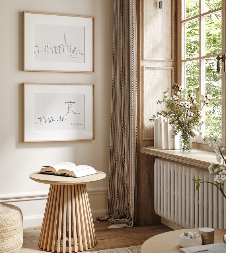 May include: Two framed black and white line drawings of city skylines, one of Dubai and one of Rio de Janeiro, hang on a beige wall. A wooden side table with a book on top sits in front of the drawings. The table is in a room with a window and a white radiator.