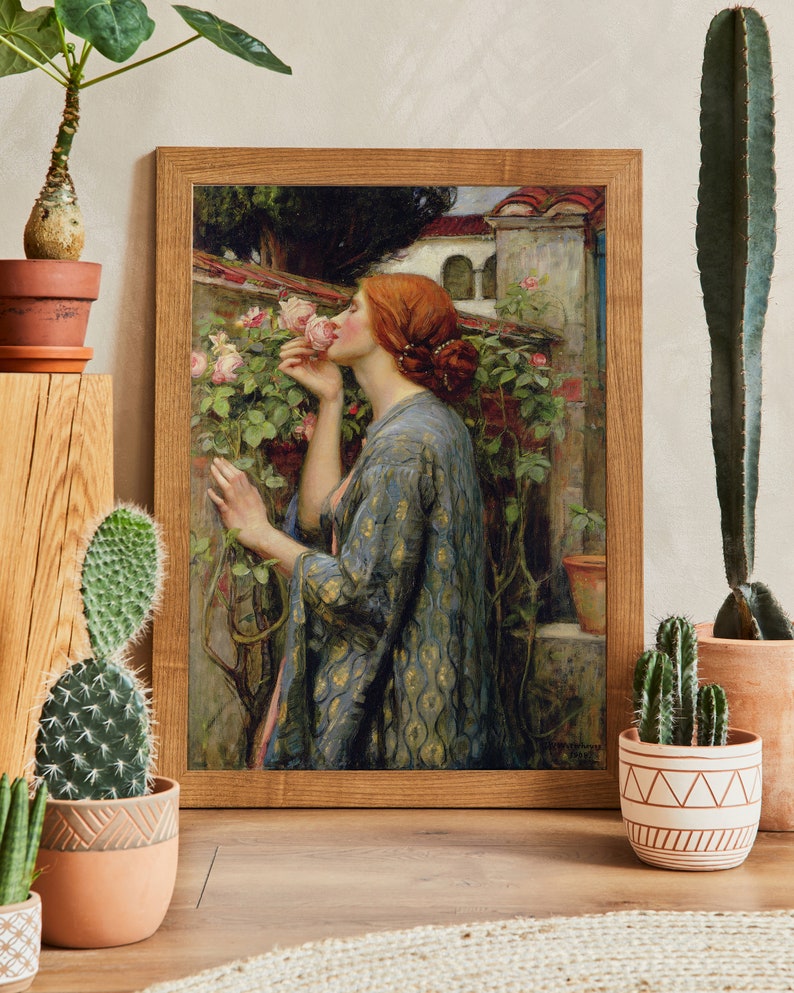 Op de afbeelding: Een ingelijst schilderij van een vrouw in een blauwe en gouden jurk die aan een roze roos ruikt. De vrouw staat in een tuin met een stenen muur en een gebouw op de achtergrond.