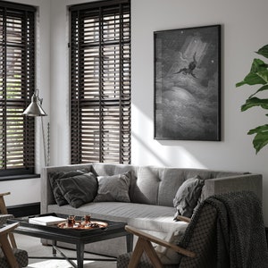 May include: A gray sofa with dark gray pillows sits in a living room with a black coffee table and a framed black and white print of a winged figure flying through the clouds. The room has brown wood blinds on the windows and a wooden chair with a black and white patterned throw blanket.