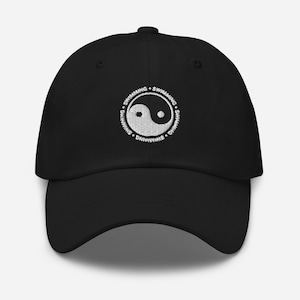 Op de afbeelding: Zwarte baseballcap met een wit geborduurd Yin en Yang-symbool, omringd door het woord "SWIMMING". De cap heeft een gebogen klep en een stoffen bandsluiting.