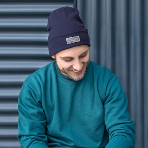 May include: Navy blue beanie with the word "MUNI" in a silver, wavy font. The beanie is ribbed and has a folded cuff. The person in the image is wearing a teal sweatshirt.