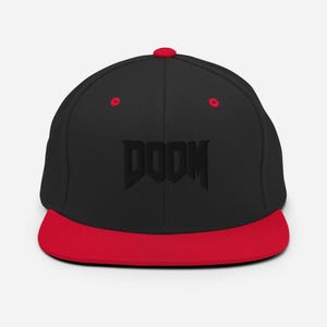 Puede incluir: Gorra de béisbol negra y roja con visera plana. La palabra "DOOM" está bordada en negro en la parte delantera. La visera, el botón superior y los ojales también son rojos.