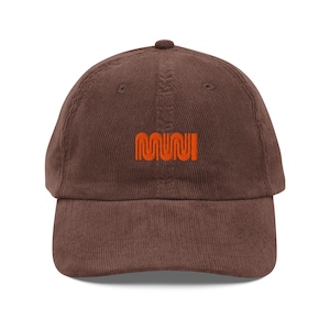 May include: A brown corduroy baseball cap with the word "MUNI" embroidered in orange thread across the front. The cap has a curved brim and an adjustable strap at the back.