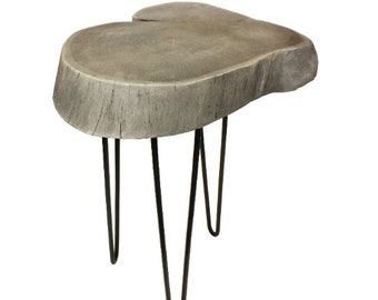 May include: A small, gray, wooden side table with a natural, organic shape. The tabletop is supported by three black, hairpin legs. The table is made of a cross-section of a tree trunk.