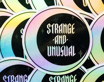 May include: A close-up of several iridescent stickers with the text "Strange and Unusual" in a black and white gothic font. The stickers have a rainbow holographic finish.