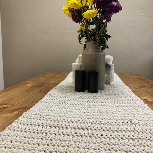 Puede incluir: Un camino de mesa de crochet beige sobre una mesa de madera con un jarrón de flores amarillas y moradas y dos candelabros negros.