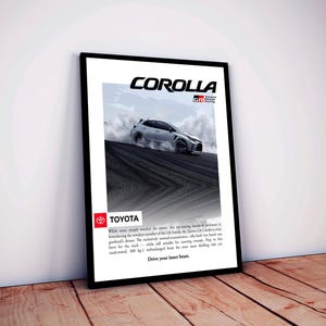 May include: Framed poster showing a white Toyota Corolla GR racing on a track, with the word "COROLLA" at the top. The Toyota logo is visible, along with text describing the car's features. The poster is leaning against a white wall on a wooden floor.