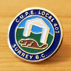 May include: A gold-coloured pin with a blue and green enamel design. The pin features a white silhouette of a building with a beaver in front of it. The text "C.U.P.E. LOCAL 402 SURREY B.C." is written around the edge of the pin.