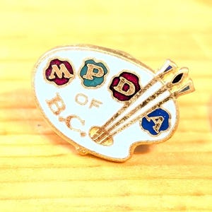 Może przedstawiać: Złoty pin w kształcie palety artysty z literami "MPDA OF B.C." w czerwonej, zielonej, niebieskiej i białej emalii. Na palecie znajdują się dwa złote pędzle. Srebrna moneta leży obok przypinki na żółtej drewnianej powierzchni.