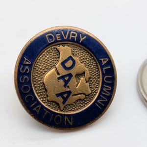 May include: A round, gold-toned DeVry Alumni Association pin with blue lettering and a blue border. The pin features the letters DAA in a gold-toned center. A coin is next to the pin.