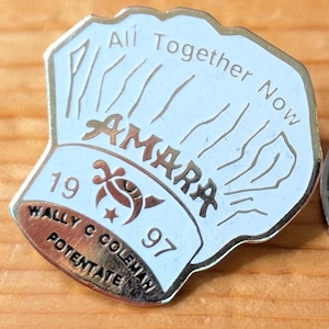 以下が含まれることがあります： 「All Together Now」と「AMARA 1997」の文字が入った銀色のコック帽ピンがコインの隣に表示されています。ピンは白い背景に銀色のトリムと、様式化されたコック帽のデザインが施されています。コインにはヘラジカが描かれています。