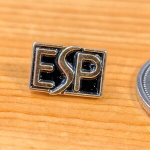 Peut inclure: Gros plan d'une épingle de revers dorée, d'une épingle argentée avec les lettres "ESP" et d'une pièce de 25 cents canadienne. L'épingle "ESP" est noire avec des lettres argentées. La pièce représente un orignal.