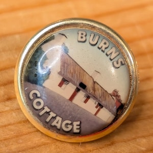 May include: A round, gold-toned pin featuring an image of a building with the text "BURNS COTTAGE". A silver coin is next to the pin on a wooden surface. The pin has a glossy finish.