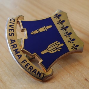 Pode incluir: Um broche dourado com um escudo azul que apresenta um canhão estilizado e uma mão segurando uma tocha. O escudo é rodeado por um rolo dourado com o texto "CIVES ARMA FERANT".