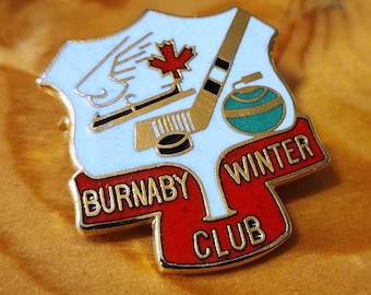 May include: A gold-colored pin with a white background featuring a hockey stick, a puck, a curling stone, and a maple leaf. The pin has red and white text that reads "Burnaby Winter Club".