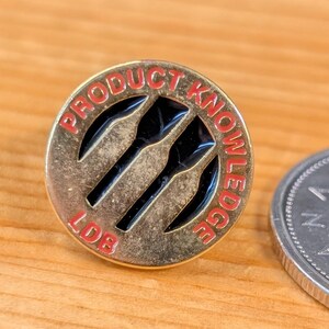 May include: A round, gold-colored pin with the words "PRODUCT KNOWLEDGE" in red lettering around the edge. The pin features three black dart-like shapes in the center. A Canadian quarter coin is next to the pin.