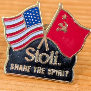 以下が含まれることがあります： アメリカとソビエト連邦の国旗が描かれたヴィンテージエナメルピン。ピンは金色のトリムで、黒い背景に「Stoli. Share the Spirit」の文字が書かれています。ピンの隣にはコインがあります。