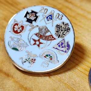 May include: A silver pin with a white enamel background and a red star in the centre. The pin features various Masonic symbols and the text "2003" and "J. BEKAL".