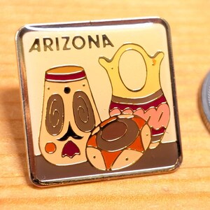 Pode incluir: Um alfinete quadrado esmaltado com a palavra "ARIZONA" acima de um desenho de três vasos de cerâmica estilizados em tons de bege, castanho, laranja e rosa. Uma moeda de prata está ao lado do alfinete em uma superfície de madeira.