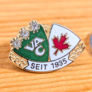 May include: A vintage enamel pin with a green and white shield design, featuring a red maple leaf and the text "SEIT 1935." The pin has gold-coloured accents and small floral details. A silver coin is next to the pin.