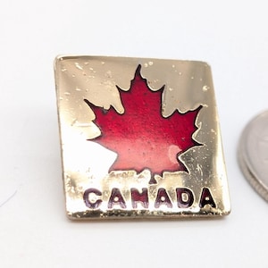 May include: A square gold-tone metal pin featuring a red enamel maple leaf at the center and the word CANADA printed in red capital letters at the bottom, positioned next to a silver-colored coin for scale on a plain white surface.