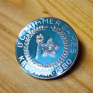 Puede incluir: Un pin de plata con un diseño azul y blanco. El pin presenta una corona de hojas, una antorcha y una flor. El texto "BC Summer Games Kelowna 1980" está escrito alrededor del borde del pin.