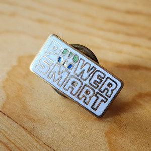 Könnte beinhalten: Eine weiße Emaille-Anstecknadel mit der Aufschrift "POWER SMART" in schwarzen Buchstaben. Die Nadel hat eine goldfarbene Metallrückseite.