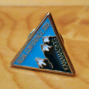 Pode incluir: Um alfinete de metal triangular com fundo azul e um design de cordilheira de montanhas brancas. O texto "RECA CONFERENCE 1985" está impresso na parte superior do alfinete e "KANANASKIS" está impresso no lado direito.