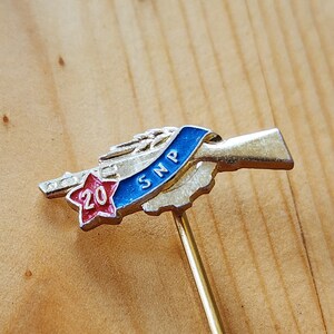 Pode incluir: Um alfinete de ouro com uma estrela vermelha e o número 20, uma fita azul com as letras "SMP" e uma imagem estilizada de um rifle.