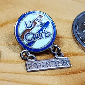 Peut inclure: Une épingle en argent avec un cercle en émail bleu, blanc et marron qui dit "U.S. Club" et une barre en argent qui dit "Founder".